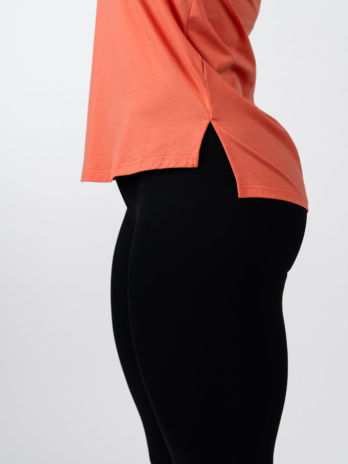Close-up side view of a person wearing a coral Women's Signature Tank paired with black leggings, highlighting the tank's soft fabric and side slit detail for ease of movement.