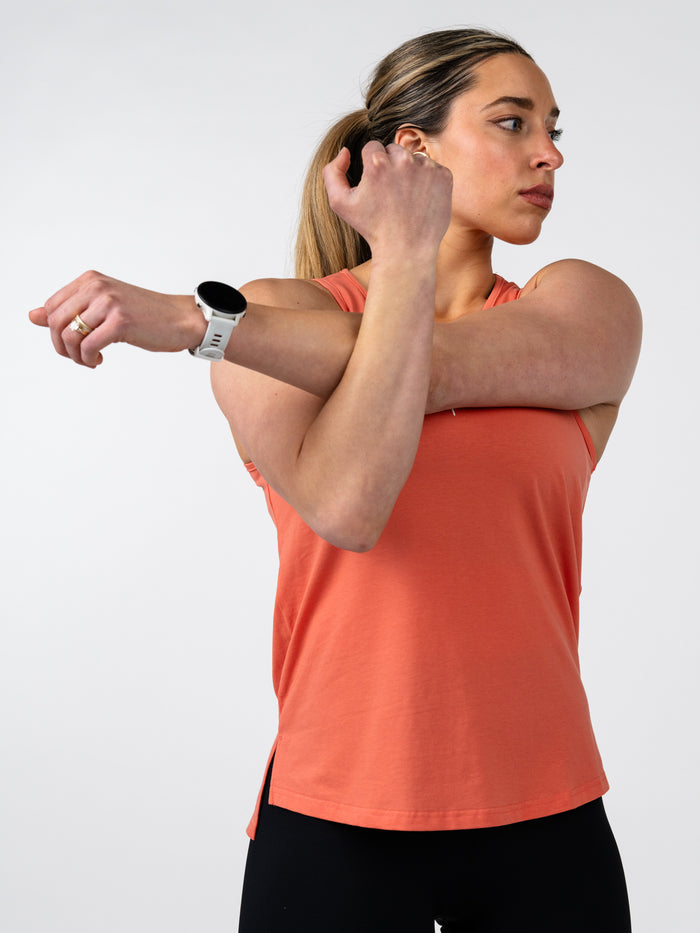 Woman wearing a coral Women's Signature Tank stretching her arm across her chest, showcasing the soft, comfortable fit ideal for activewear and fitness activities.
