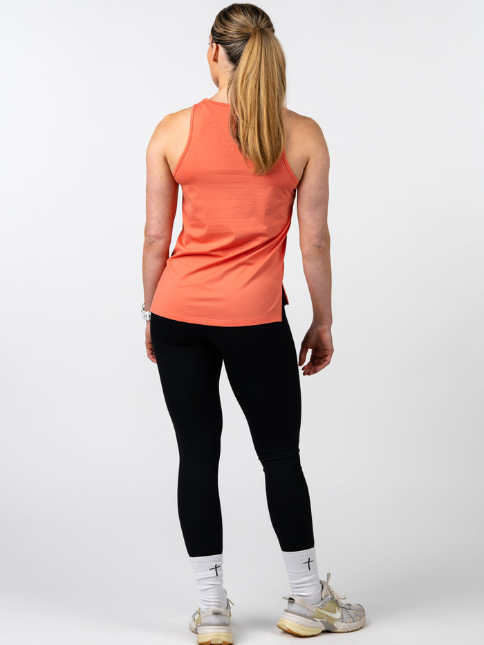 Woman wearing a coral Women's Signature Tank paired with black leggings and white socks with a cross design, standing facing away on a plain background.